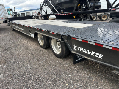 Trailer shows a long flatbed with diamond plate surface and dual rear axles. Side branding reads TRAIL-EZE with a white circular logo. Elevated equipment and black paint with red/white reflective tape along edge.