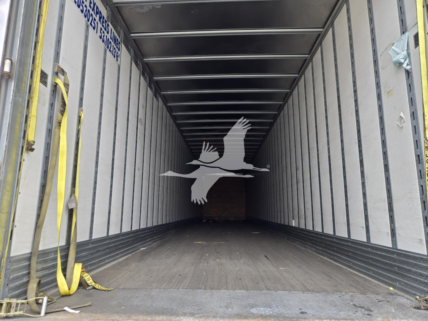 Inside cargo trailer showing empty V-shaped interior, wooden floor, metal side walls, yellow straps along left wall.