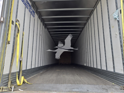 Inside cargo trailer showing empty V-shaped interior, wooden floor, metal side walls, yellow straps along left wall.