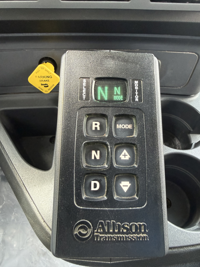 Allison transmission gear selector with lights showing N and M mode, adjacent are R, N, D, plus and minus buttons; yellow parking brake knob on left; cup holders visible.