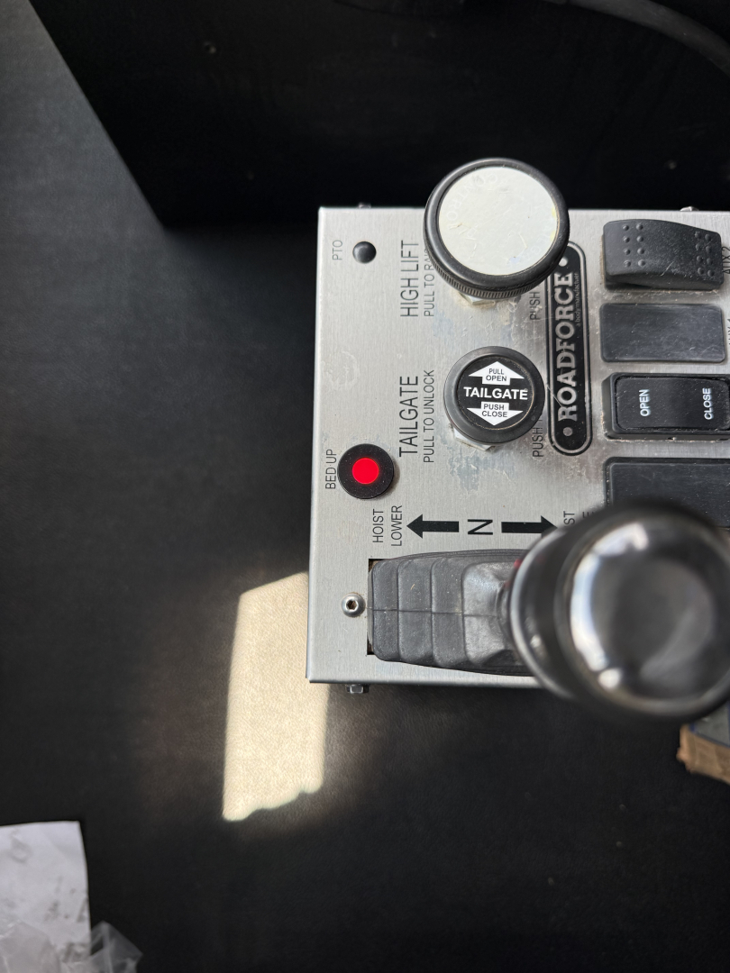 Dashboard control panel in a Kenworth T880S cab showing a toggle lever, push buttons, and labels such as ‘TOOL’, ‘GATE’, and ‘TAYL-…’ with a circular gauge knob and switch; close-up view.