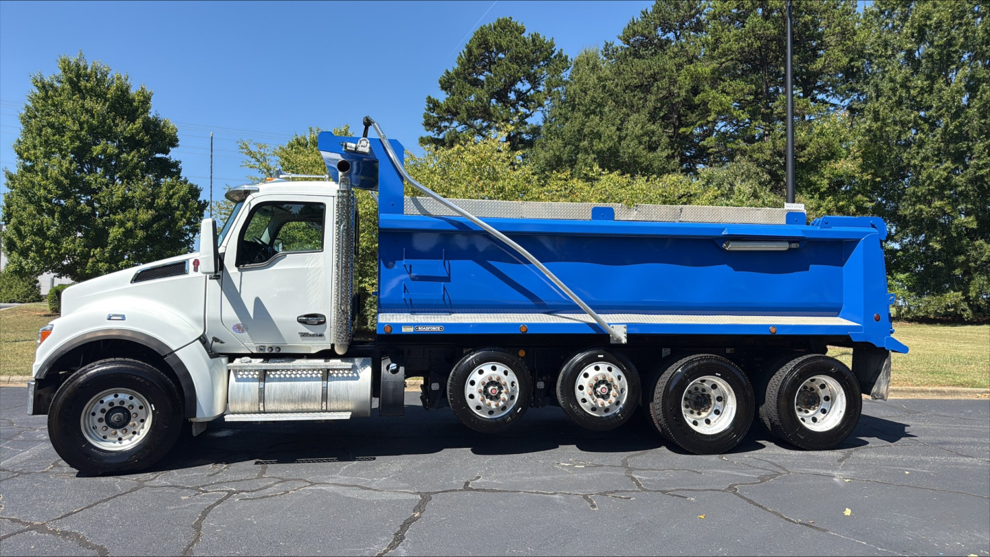 Dump truck showing 2024 Kenworth T880S with blue dump body and white cab, side profile view.