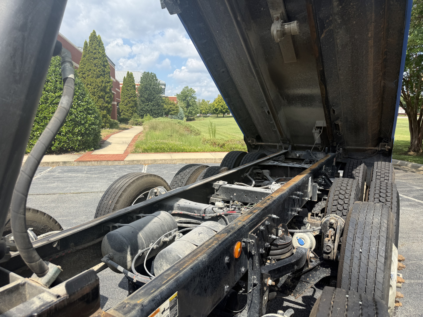 Truck chassis in view with raised dump bed showing frame, suspension, and dual rear wheels.