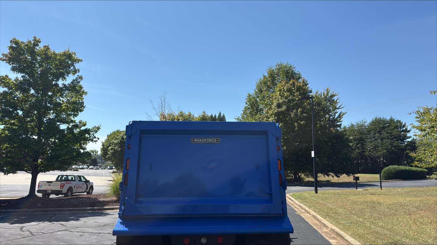 Blue Kenworth T880S dump truck bed with rear view, showing a rectangular blue tailgate and surrounding trim; a white vehicle and trees in the background.