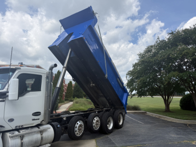 Side view of a white Kenworth truck with a raised blue dump bed in a parking lot, revealing multiple rear wheels and hydraulic lifting mechanism.