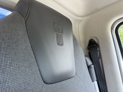 Kenworth logo on seat backrest in truck cabin, with seatbelt and interior panel in view.
