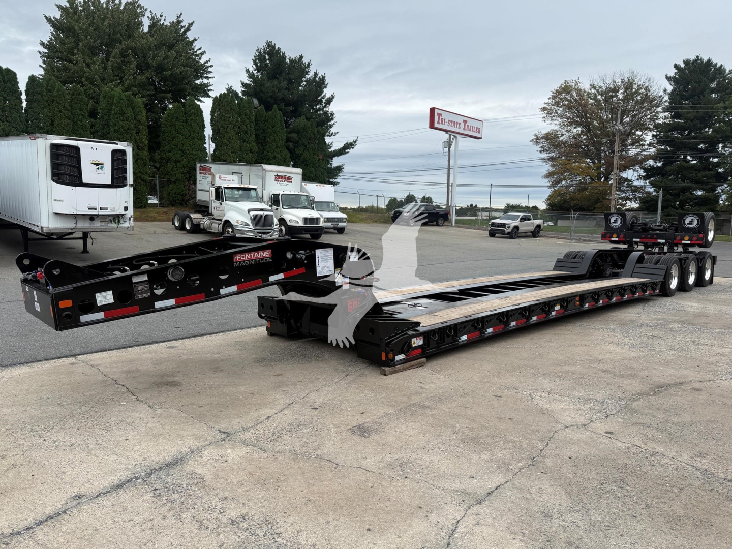 Lowboy trailer setup showing Fontaine Magnitude 60Lcc label and dual long black lowbed with multiple axles, reflective tape, and adjacent equipment in a dealer lot.