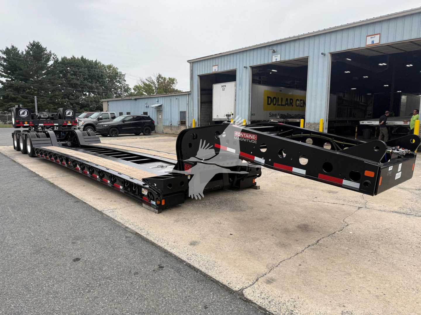 Lowboy trailer with Fontaine Magnitude 60Lcc plate and gooseneck, blue-gray shop in background.