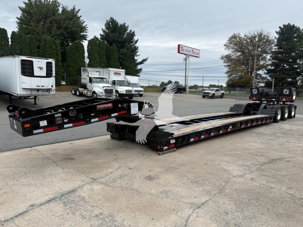 Lowboy trailer setup showing Fontaine Magnitude 60Lcc label and dual long black lowbed with multiple axles, reflective tape, and adjacent equipment in a dealer lot.