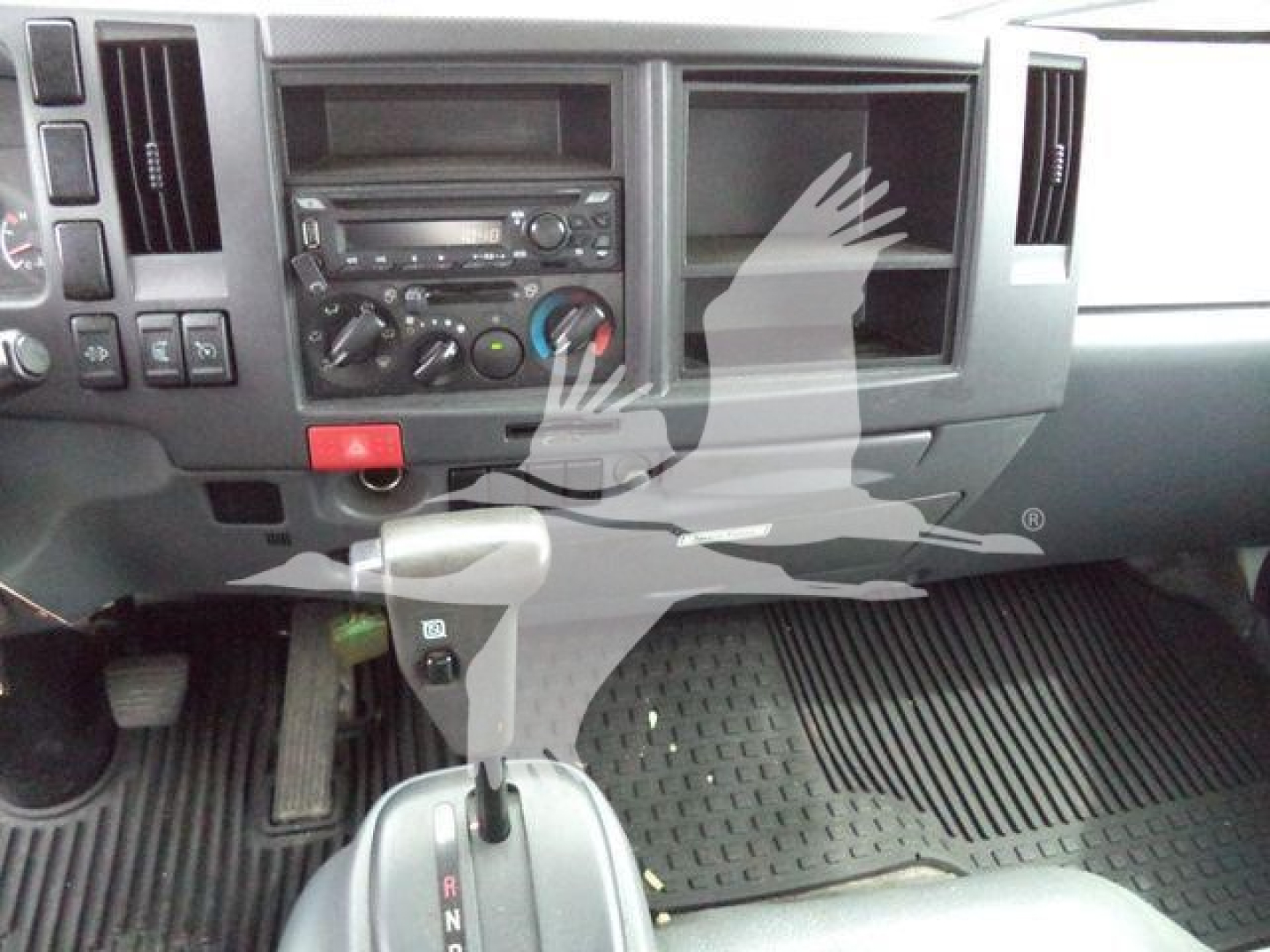 Isuzu NPR/NRR cab interior showing center dashboard with radio, climate controls, and gear shifter; matte black floor mats; empty passenger side dash pocket.