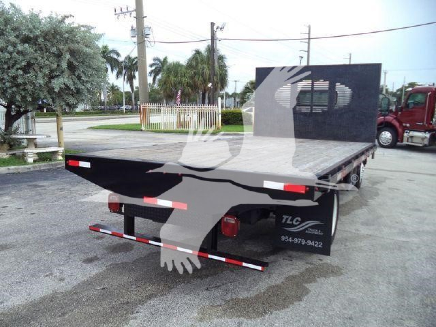 Isuzu NPR flatbed truck rear view showing flatbed deck and TLC branding on side; tail lights and rear bumper visible.