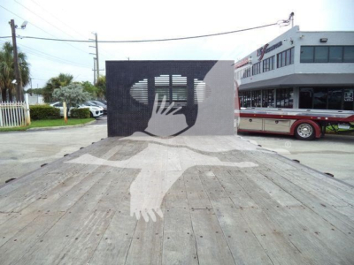 Flatbed tow truck with wooden surface and a metal ramp featuring a cutout hands design; dealership building in background.
