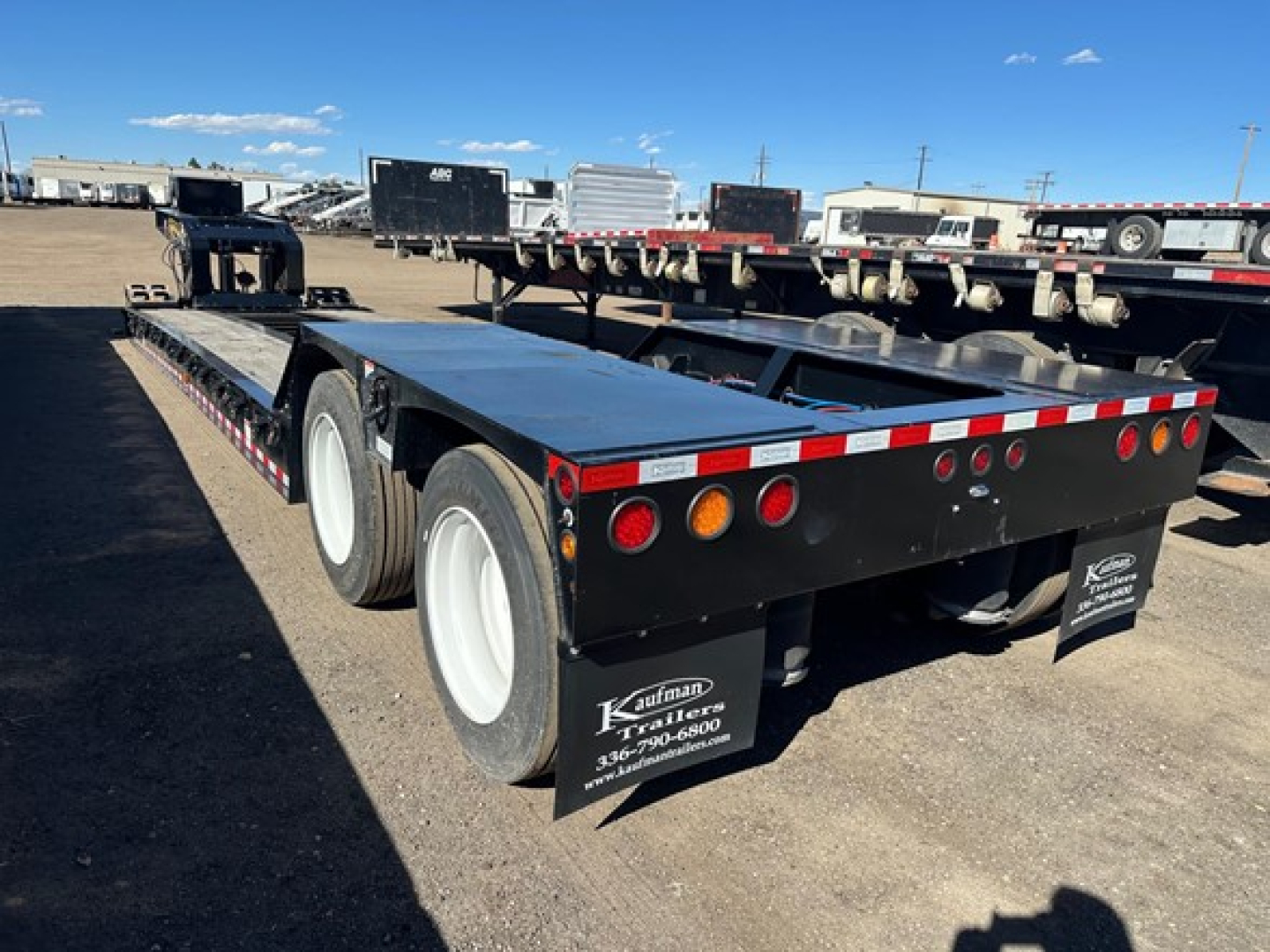 Lowboy trailer showing dual rear axles and flatbed deck with black side panels and round taillights.