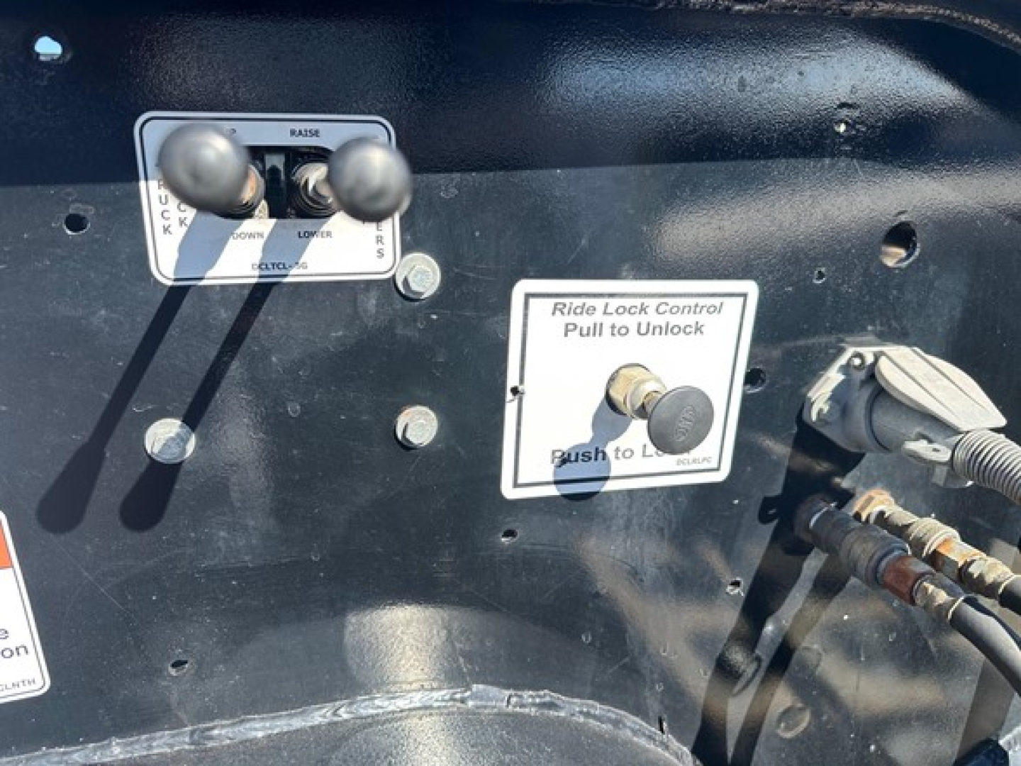 Lowboy trailer control panel with two black levers, labels reading Ride Lock Control: Pull to Unlock, Push to Lock, and other warning stickers and hoses visible.