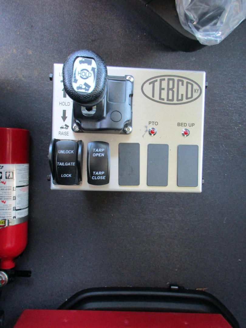 Cab control panel with TEBCD logo and joystick-like lever; labels 'PTO', 'BED UP', 'TARP OPEN', 'TARP CLOSE', warning LEDs, and small black buttons, mounted on a gray panel beside a red fire extinguisher.