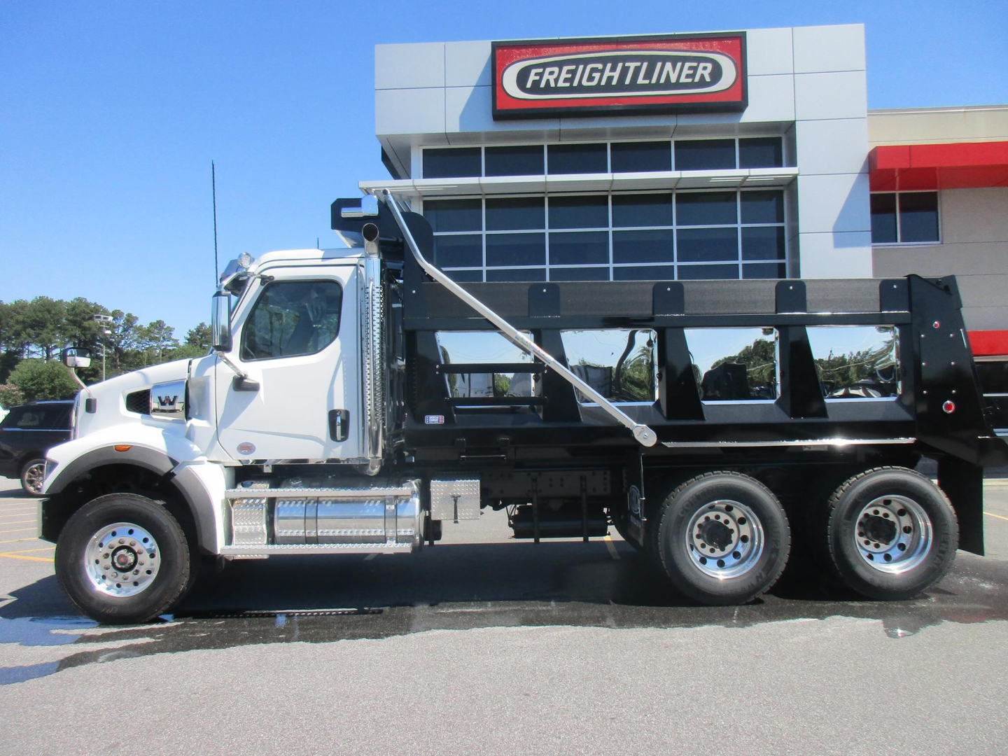 New Western Star 47X dump truck with white cab and black dump body parked in front of a Freightliner dealership building.