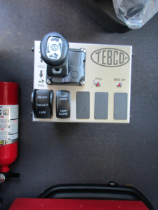 Cab control panel with TEBCD logo and joystick-like lever; labels 'PTO', 'BED UP', 'TARP OPEN', 'TARP CLOSE', warning LEDs, and small black buttons, mounted on a gray panel beside a red fire extinguisher.