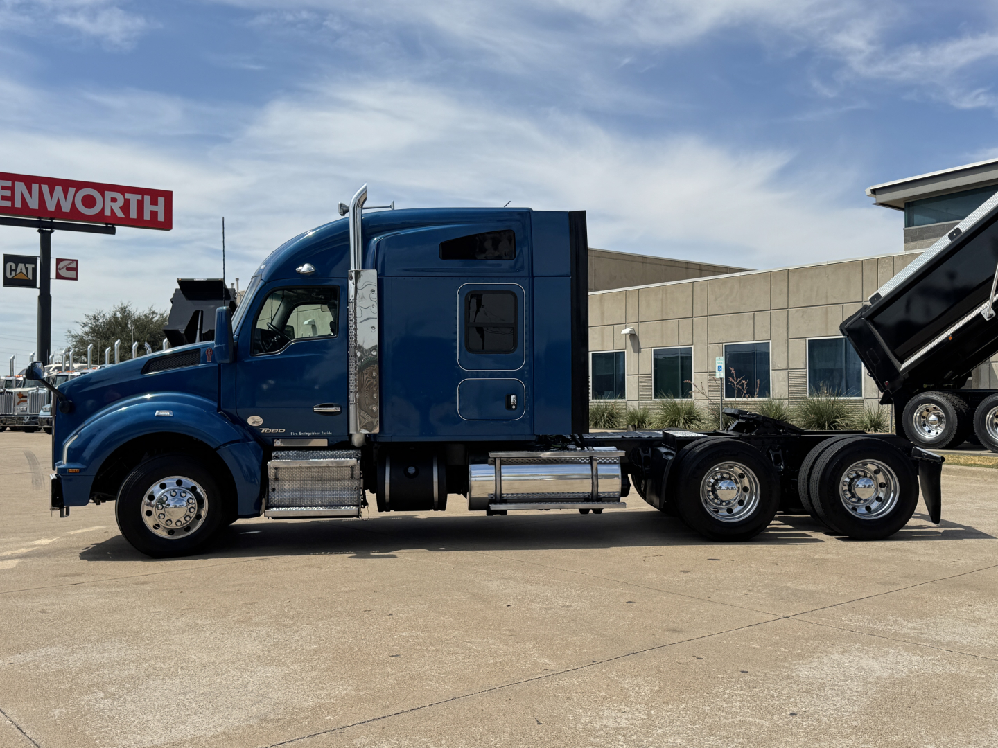 Kenworth T880 conventional sleeper tractor in blue, side profile, showing fuel tanks, steps, and dual rear axles with a dump trailer in the background.
