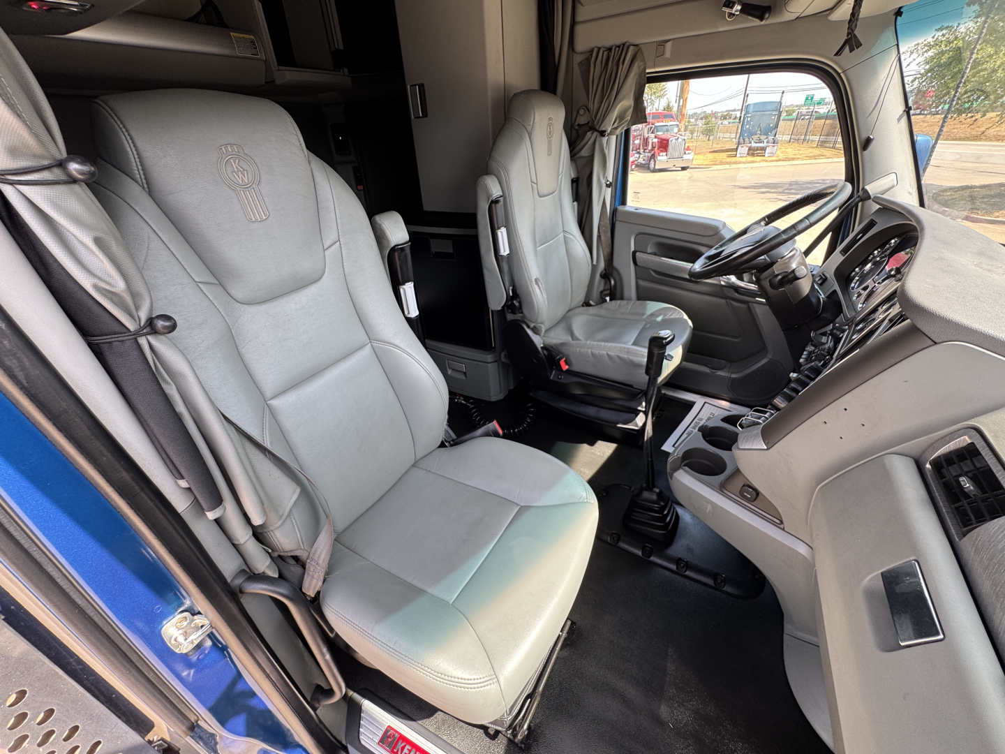 Cab interior of a 2019 Kenworth T880: driver and passenger seats, steering wheel, dashboard, gear shifter, and window view; light gray seats with embroidered logo on headrest.
