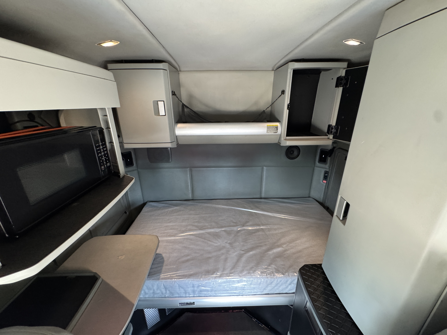 Interior cab of a 2019 Kenworth T880 sleeper with a bed, overhead storage, microwave on left, and storage compartments. Visible labels and components include a bed mattress, cabinet doors, and a small yellow caution sticker.