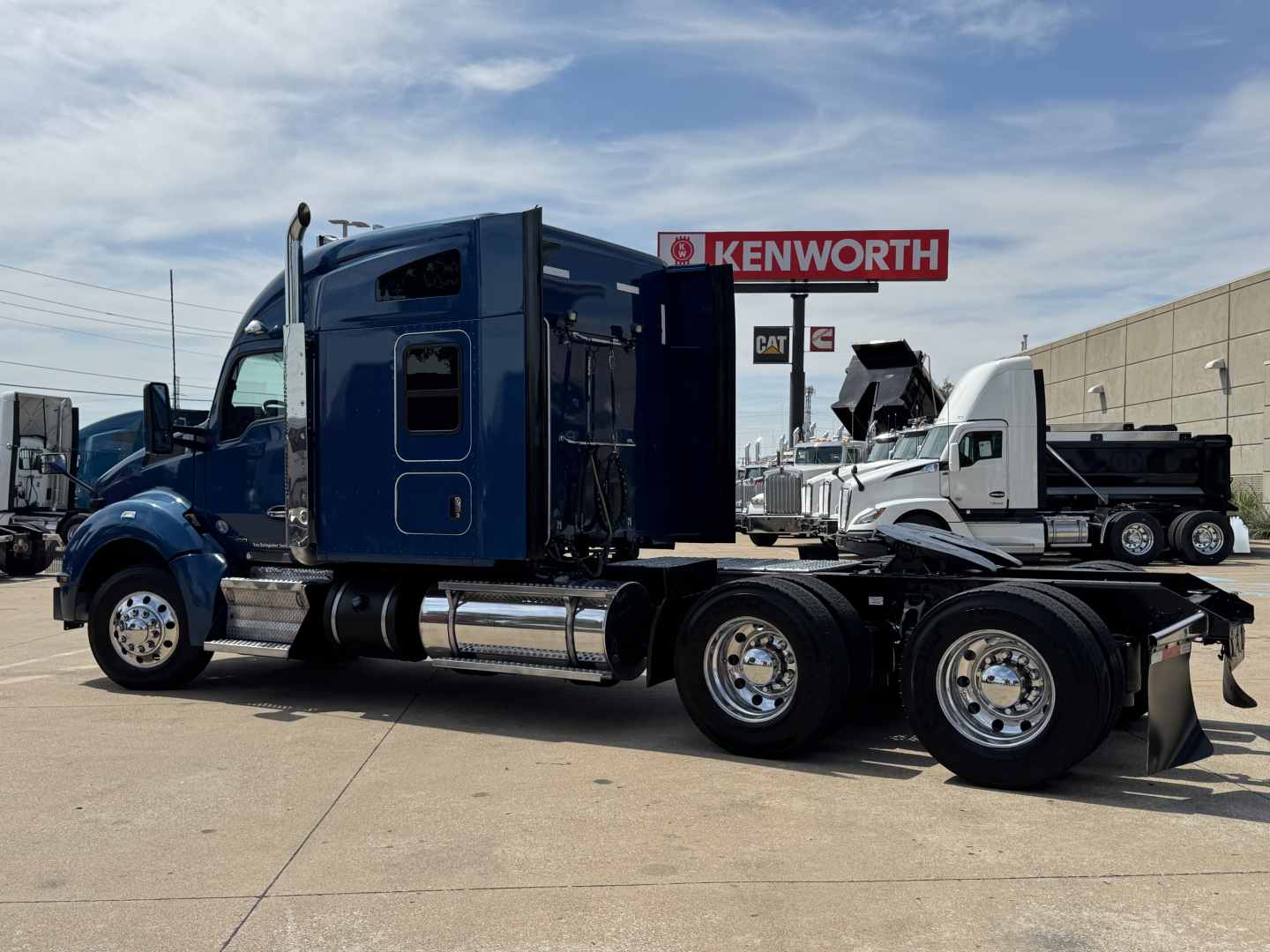 Kenworth T880 tractor with dark blue cab, side view showing sleeper, chrome exhausts, and dual rear axles. Visible text includes Kenworth dealership sign in background.