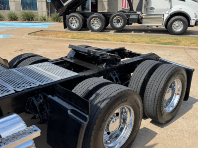 Kenworth T880 chassis and dual rear axles with large polished wheels, fifth wheel and metal steps; cab-over truck in background; daytime outdoor lot.