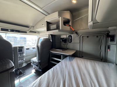 Kenworth T880 sleeper cab interior showing overhead bunk area with mounted monitor and storage compartment, driver seat, dashboard, and bed in foreground.