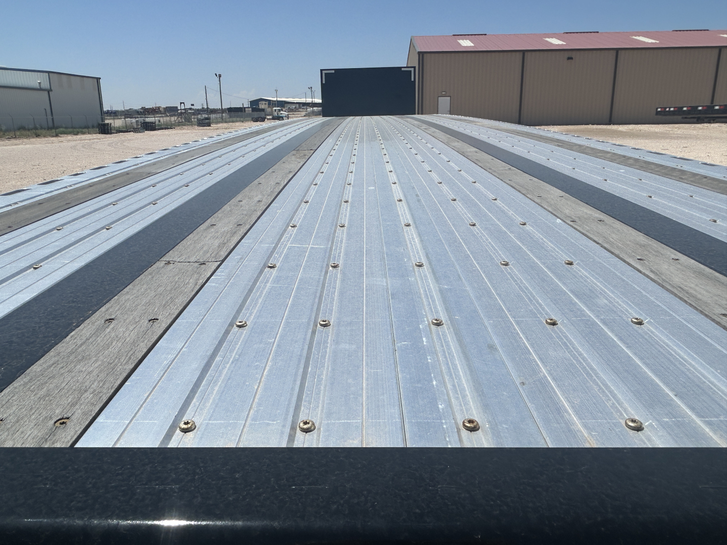 Flatbed trailer deck with metal ribs and fasteners; view extends toward structures in the distance.