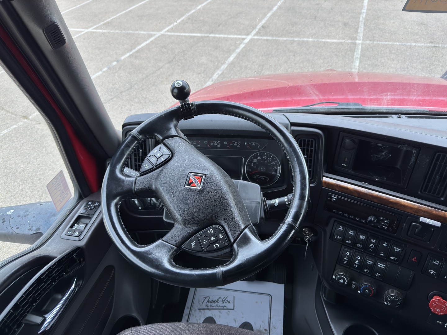 Truck interior showing steering wheel and dashboard of a red 2018 International LT625 with Clutch gear shifter on dash, various controls, and a name tag mat.