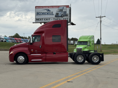2018 International LT625 sleeper tractor with red cab and green trailer unit, side view, on road with billboard in background.