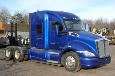 Kenworth T680 sleeper cab tractor in blue, side view showing front grille, headlights, and dual rear axles; vehicle is in a yard with other equipment.