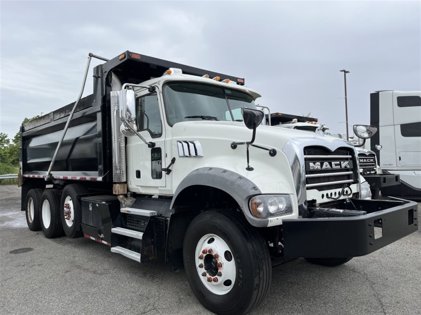 Dump truck showing Mack GR84F dump bed with white cab and black bumper, front grille with MACK branding, six wheels visible, side mirror, and hydraulic bed components.