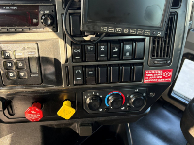 Mack GR84F cab interior showing dash controls with red and yellow hazard knobs, climate dials, and a touchscreen monitor.