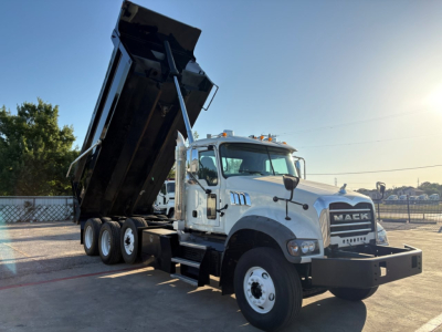 Dump truck in-service Mack GR84F with raised dump bed, white cab, black dump body, heavy-duty front bumper, dual rear axles, daylight sun, industrial lot.