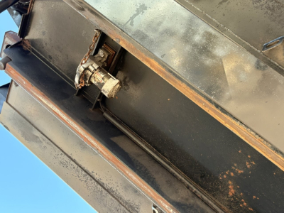 Dump truck component with hydraulic hinge and a small pneumatic/valve assembly mounted on a metal panel; visible rust and wear on the edge.