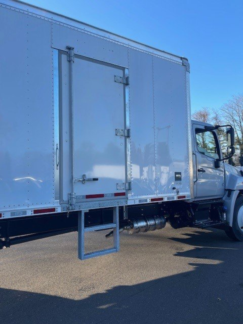 Box truck side showing entry door and step, tandem fuel tanks, cab visible on right, white exterior, clear sky.