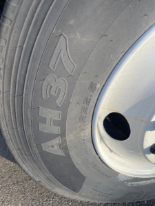 Truck wheel and tire close-up showing tire marked 'A H3' and wheel hub; part of box van truck listing for a 2026 Hino L6.