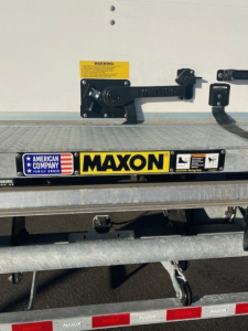 Rear of a Maxon trailer with a yellow warning sticker, latch, and Maxon sticker on the tailgate; American flag and 'MAXON' branding visible.