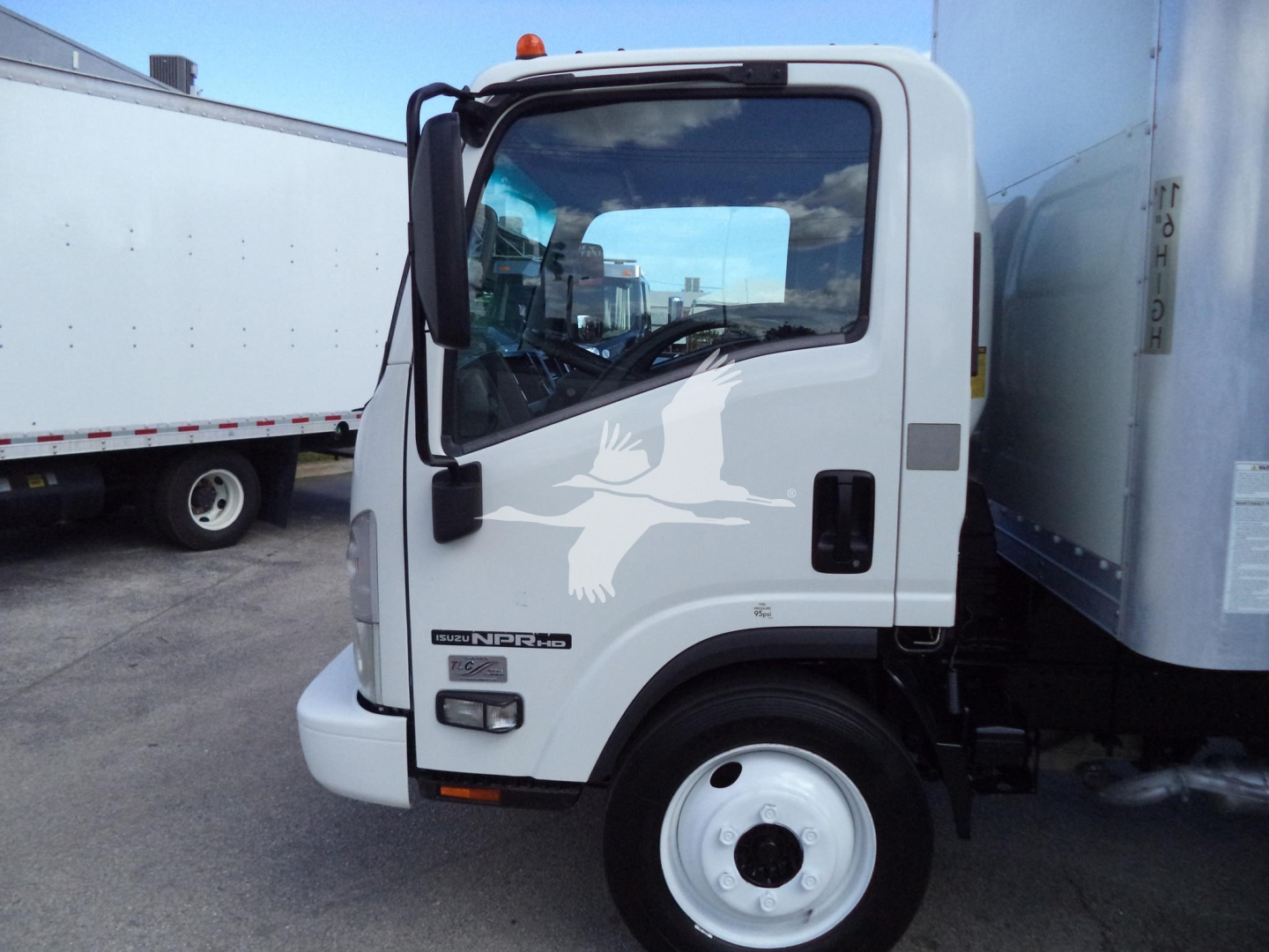 Isuzu NPR HD box truck cab front view showing side mirror, windshield, and door emblem; white cab with logo and fuel/air components visible.