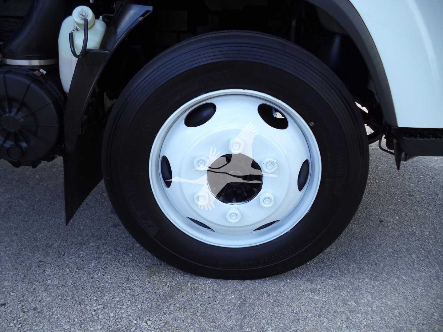Isuzu NPR HD truck front wheel and wheel well with white fender, black tire, and silver hubcap; visible brake components and reservoir nearby.