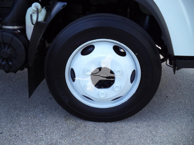 Isuzu NPR HD truck front wheel and wheel well with white fender, black tire, and silver hubcap; visible brake components and reservoir nearby.