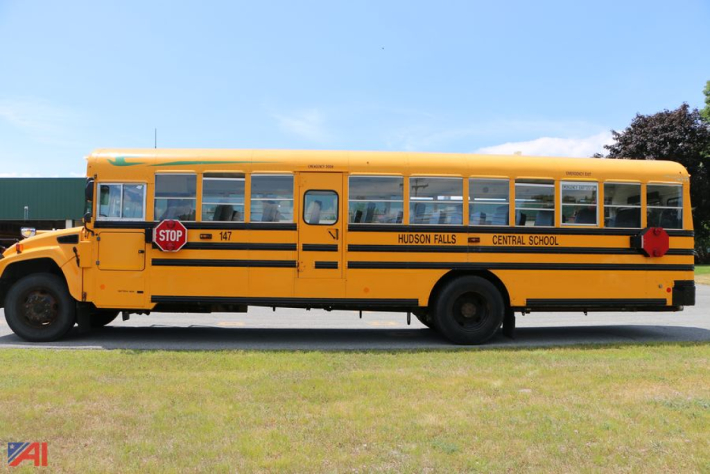 Yellow school bus side view showing 'HUDSON FALLS CENTRAL SCHOOL' lettering, black stripes, red stop signs and bus number 147.