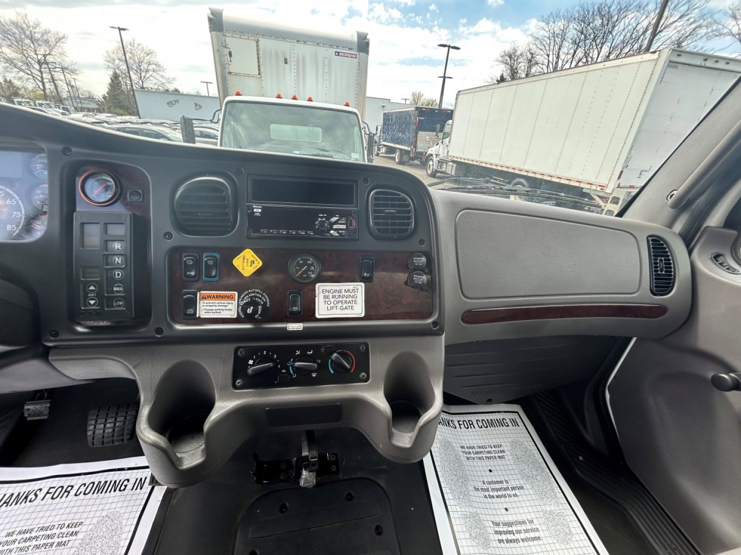 Dashboard of a Freightliner M2 106 cab showing instrument cluster, steering wheel, center control panel, and wood-grain trim; visible switches and warning labels.