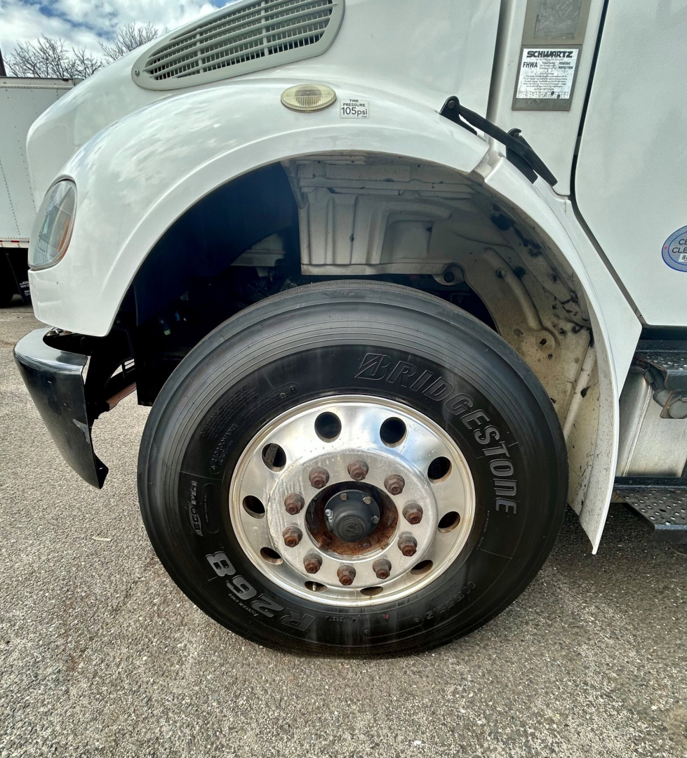 Wheel area of a Freightliner M2 106 front left showing Bridgestone tire and wheel hub, fender and side panel with badge; partial engine bay and wheel well visible.
