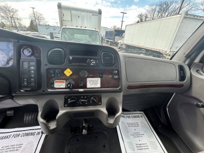 Dashboard of a Freightliner M2 106 cab showing instrument cluster, steering wheel, center control panel, and wood-grain trim; visible switches and warning labels.