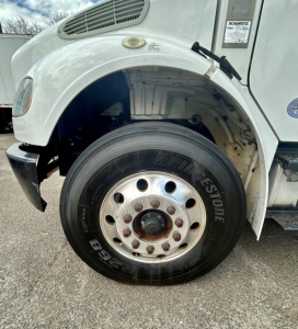 Wheel area of a Freightliner M2 106 front left showing Bridgestone tire and wheel hub, fender and side panel with badge; partial engine bay and wheel well visible.