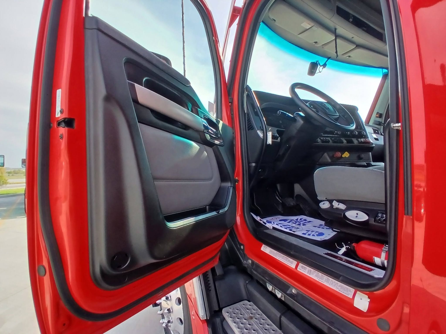Truck cab interior visible with open door showing steering wheel and dashboard from driver side. Red exterior, gray and black interior components.