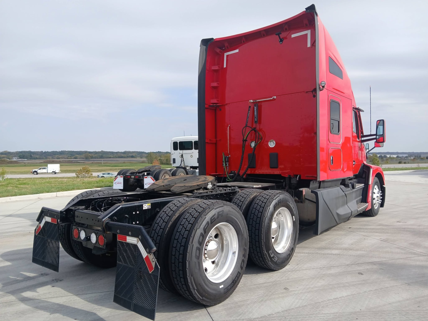 Kenworth T680 tractor in red cab, rear view showing triple rear axles, fifth wheel, and dual mud flaps; license-free lot in background.