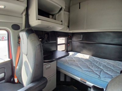 Interior of a Kenworth truck cab with a bunk bed and storage drawers; visible labels include no obvious text but the scene shows a sleeping area with a blue mattress and overhead cabinets.