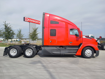 Kenworth T680 semi-truck in red with sleeper cab, side view showing dual rear axles and exhaust, Kenworth dealership sign in background.